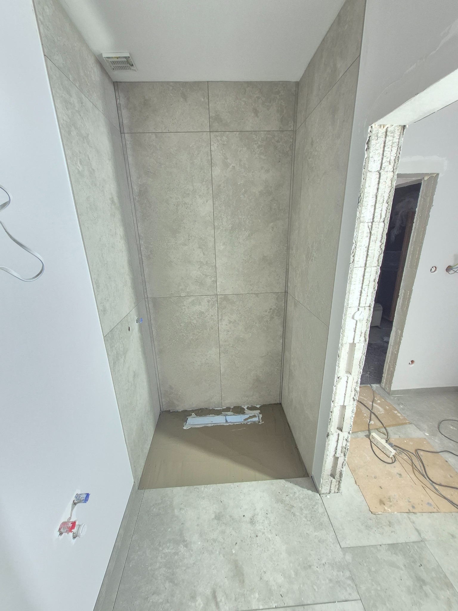 Wnętrze łazienki w trakcie wykańczania: ułożone szare płytki w kabinie prysznicowej, widoczne instalacje wodne i fragmenty niezabezpieczonych ścian.