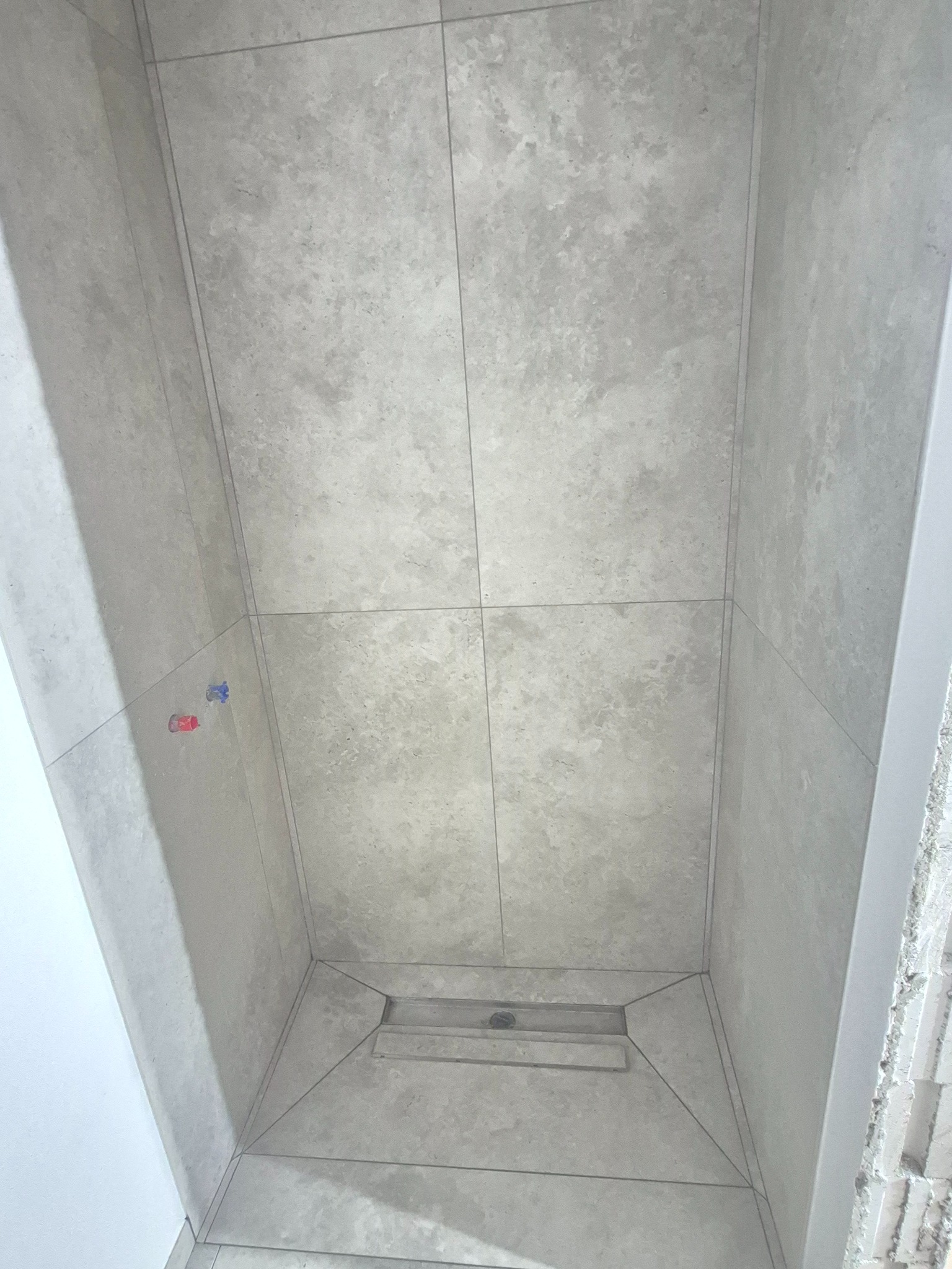 Wykończony brodzik z odpływem liniowym i szarymi płytkami imitującymi kamień. Widoczne przyłącza wodne. Minimalistyczny design.