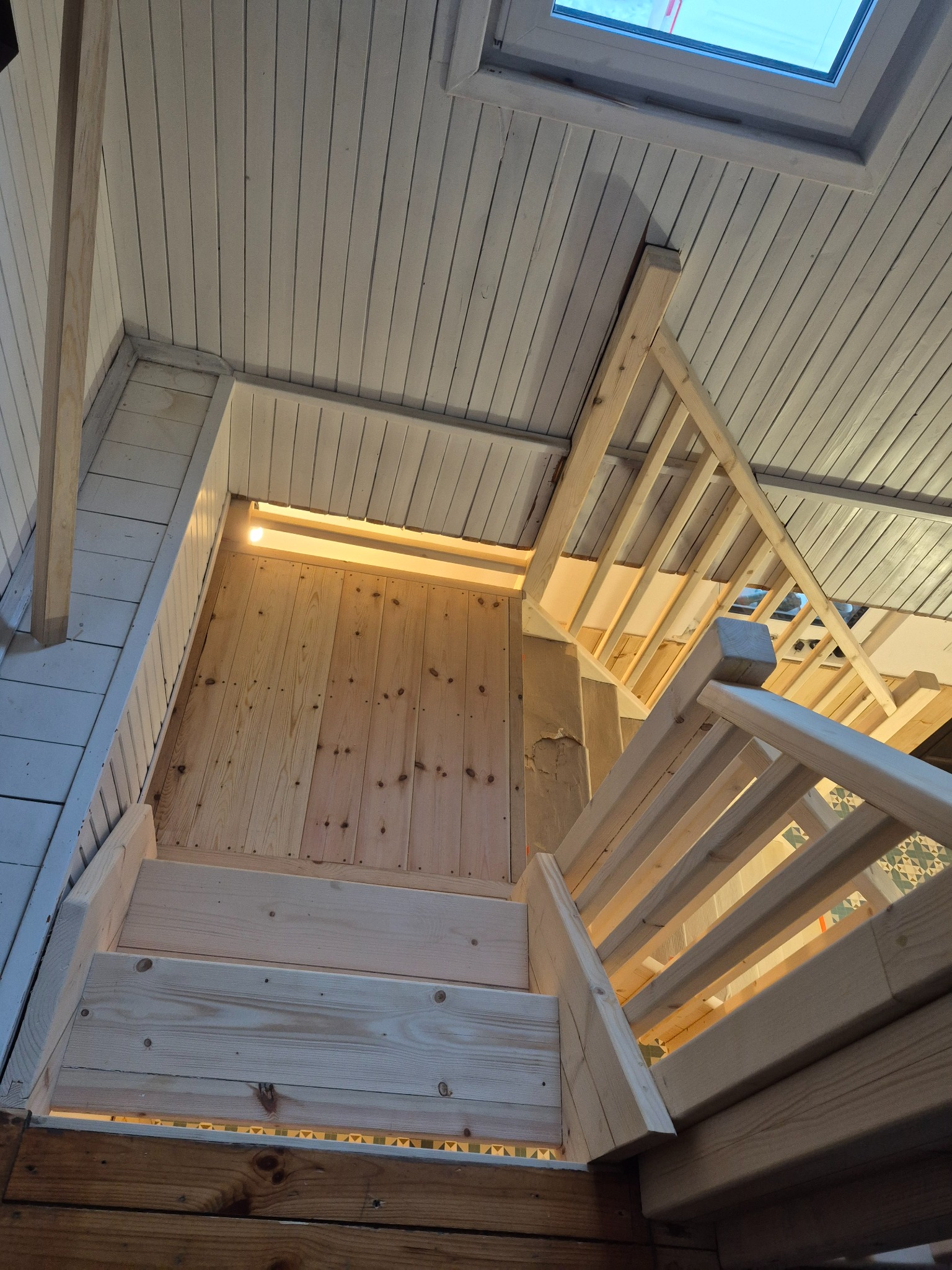 Drewniane schody z balustradą w jasnym wnętrzu z oknem dachowym. Widoczne deski na suficie i podłodze. Naturalne światło podkreśla strukturę drewna.