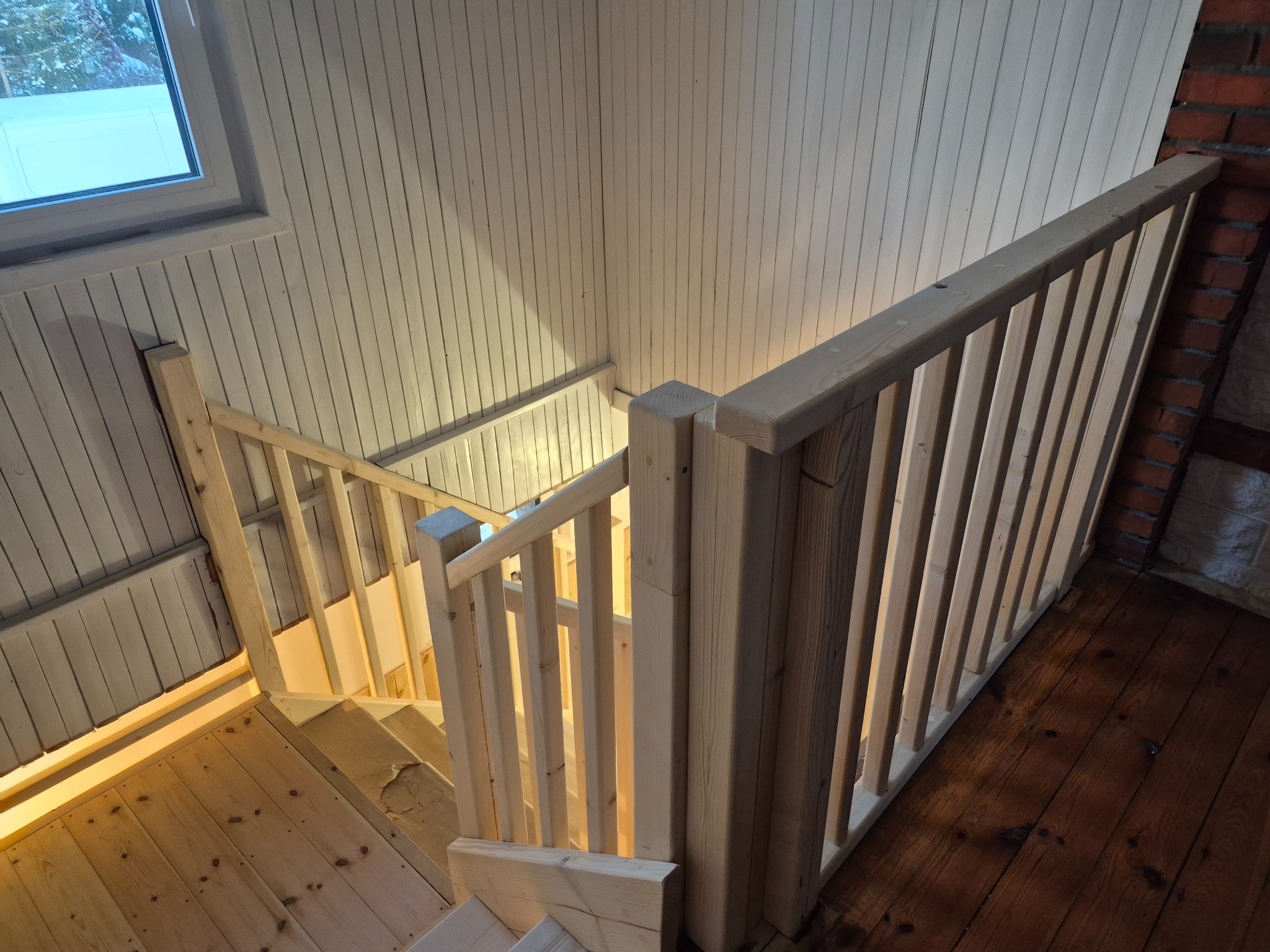 Drewniane schody z balustradą w jasnym wnętrzu. Widoczne deski podłogowe i boazeria na ścianach. Ujęcie z góry, podkreślające solidne wykonanie i naturalny charakter materiałów.