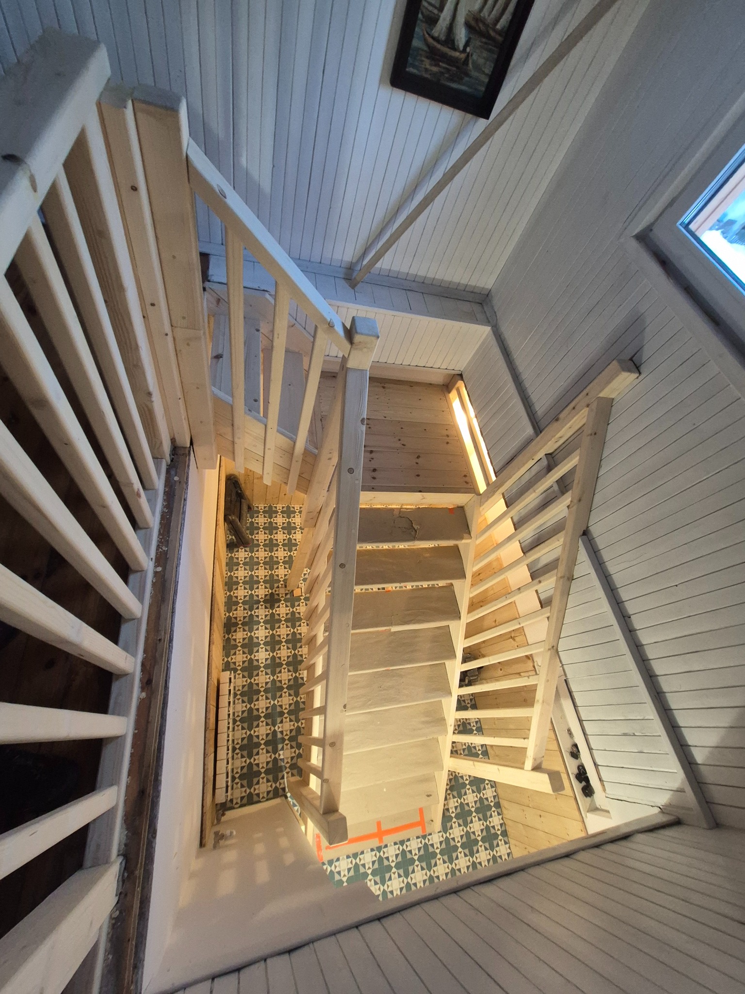 Drewniane schody z balustradą w jasnym wnętrzu z boazerią. Widok z góry ukazuje wzorzystą podłogę i obraz na ścianie. Surowe drewno kontrastuje z wykończonymi powierzchniami.