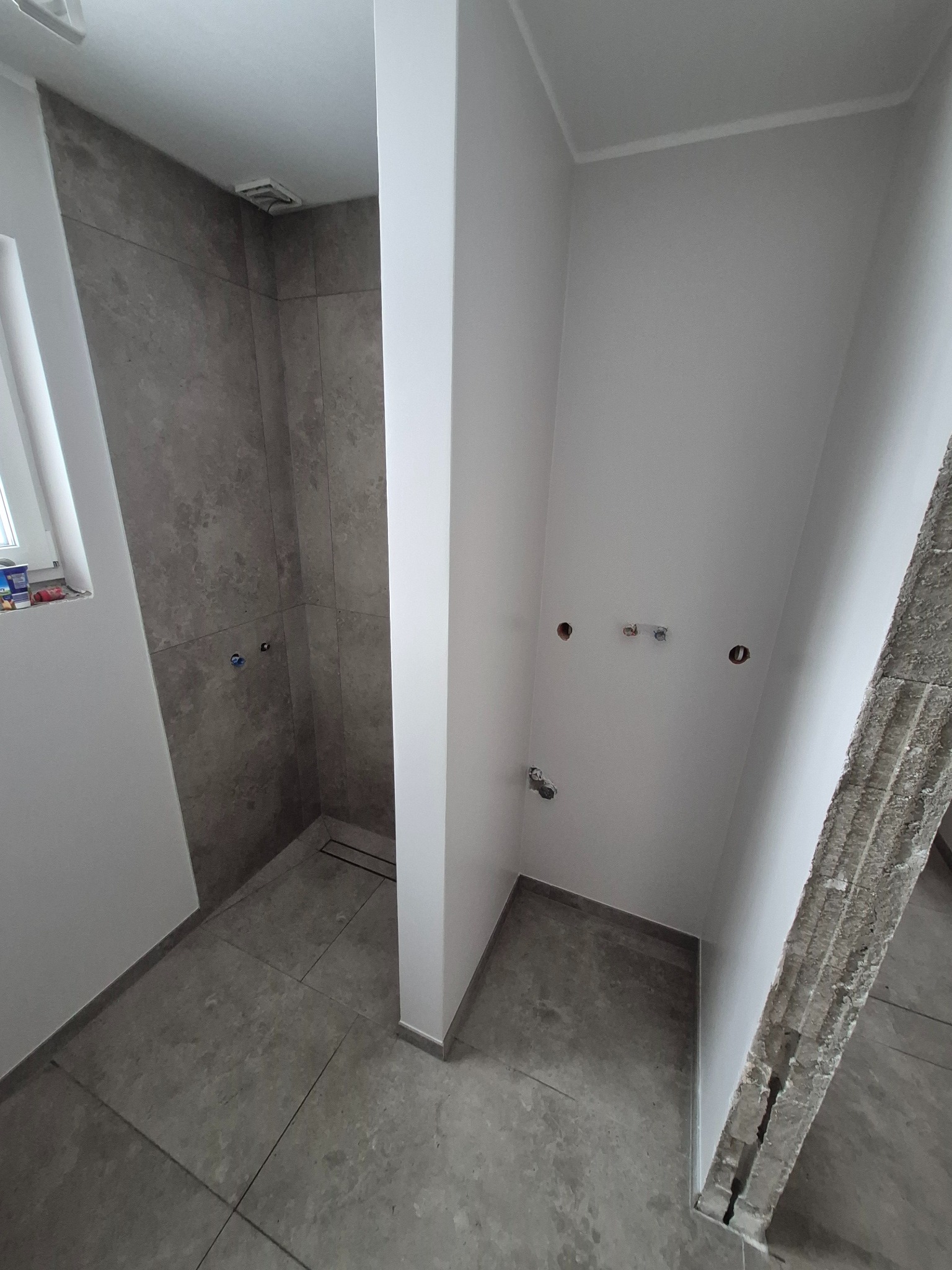 Surowa łazienka z szarą glazurą w kabinie prysznicowej i przygotowanymi punktami instalacyjnymi na białych ścianach. Widoczny fragment ściany z odsłoniętą konstrukcją.