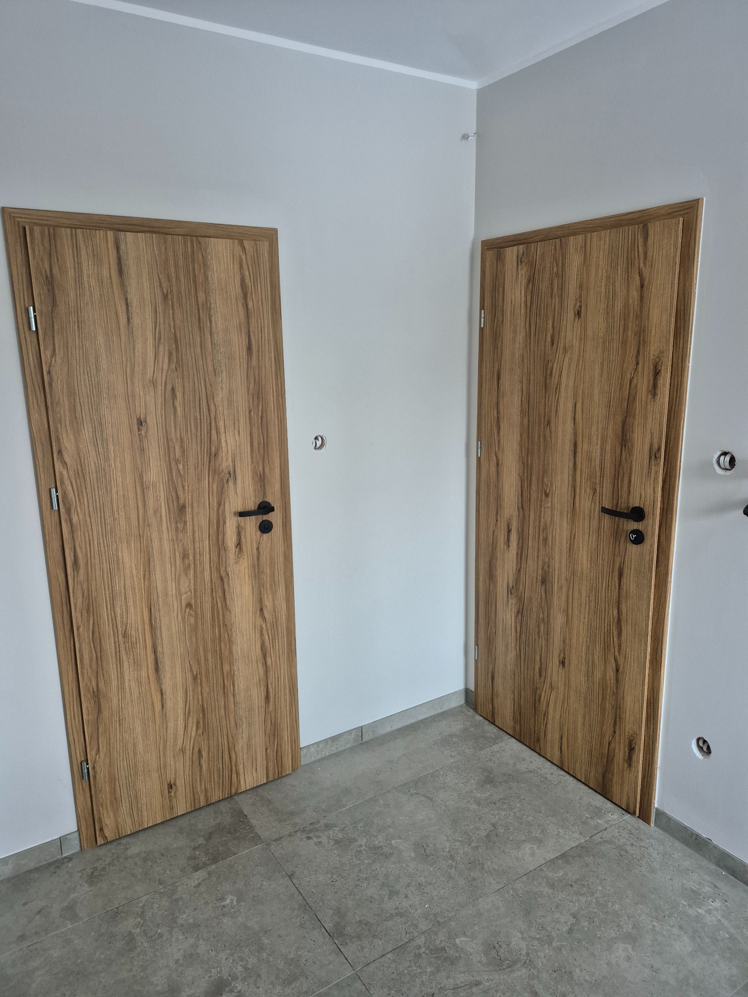 Dwie drewniane drzwi wewnętrzne z czarnymi klamkami, zamontowane w rogu pomieszczenia z szarą podłogą. Widoczne gniazdka elektryczne i otwory w ścianie.