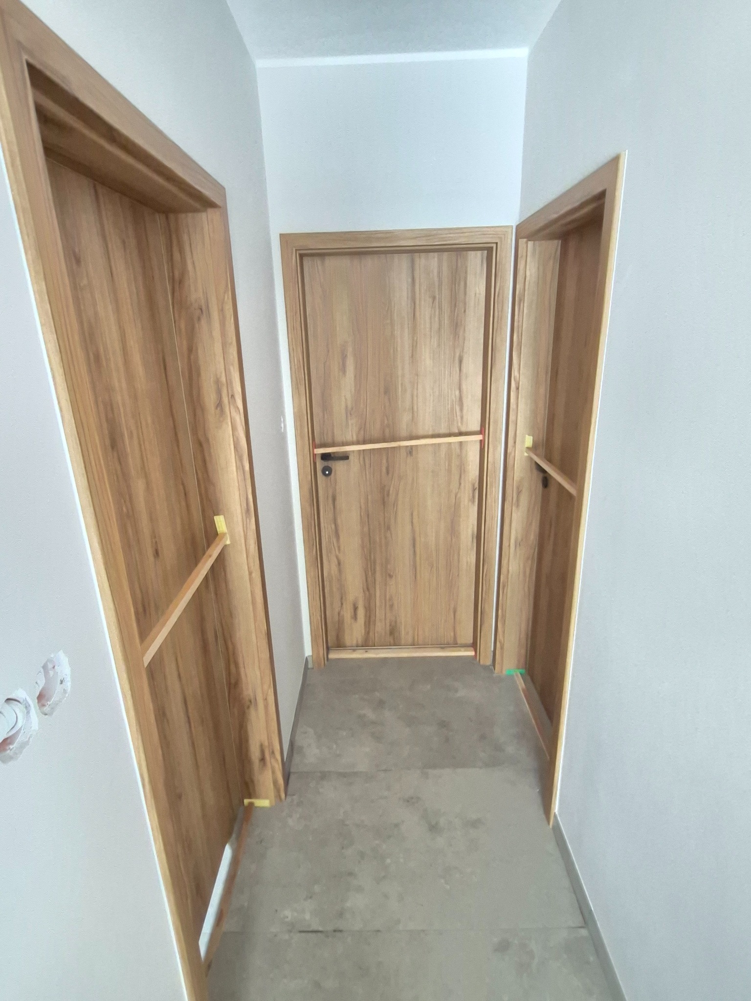 Trzy drewniane drzwi wewnętrzne z ościeżnicami, zamontowane w jasnym korytarzu. Dwa skrzydła drzwiowe są częściowo otwarte, zabezpieczone listwami montażowymi.