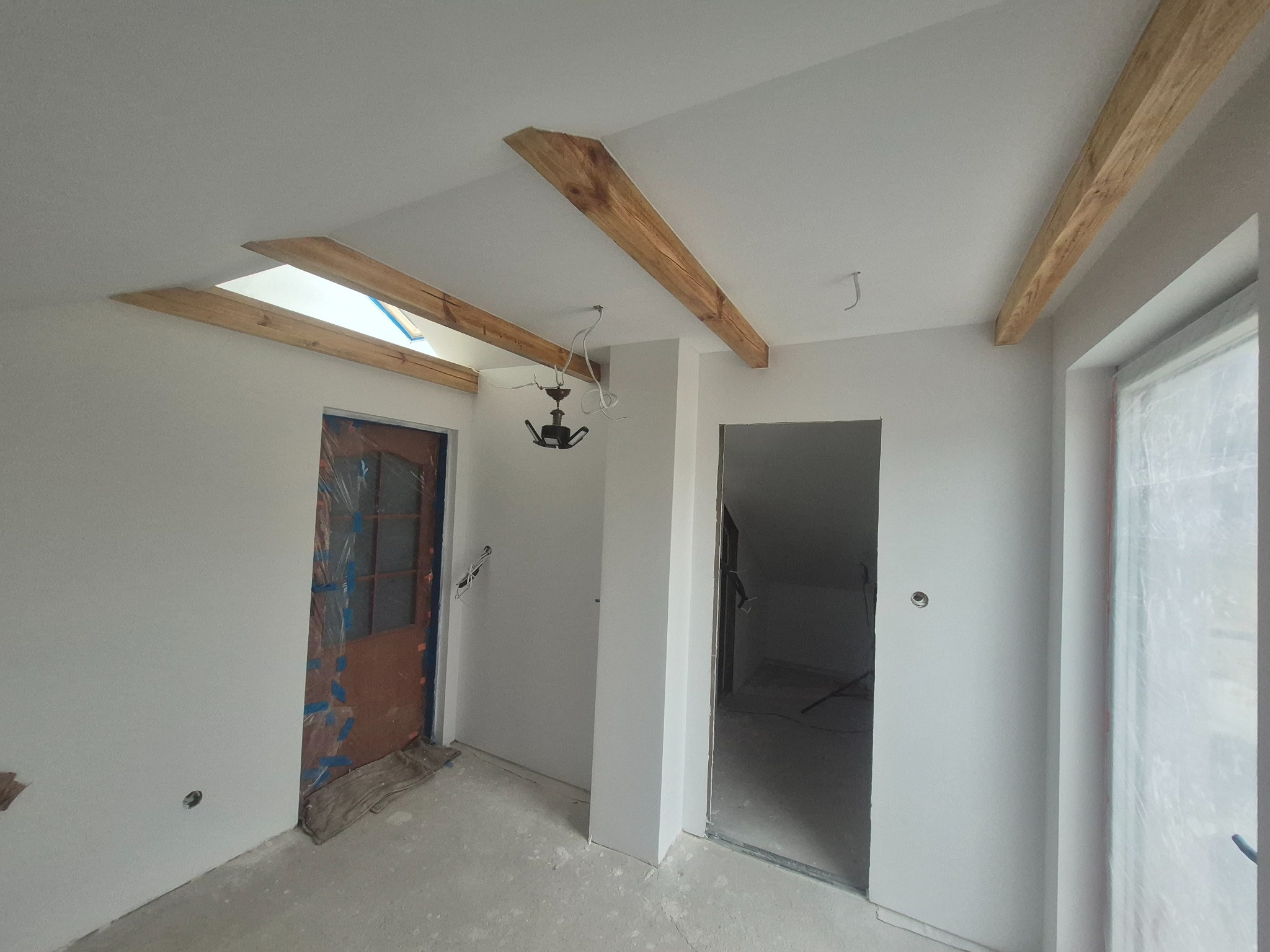 Wnętrze poddasza po malowaniu ścian na biało, z drewnianymi belkami stropowymi i oknem dachowym. Drzwi zabezpieczone folią malarską, widoczny stan po malowaniu.