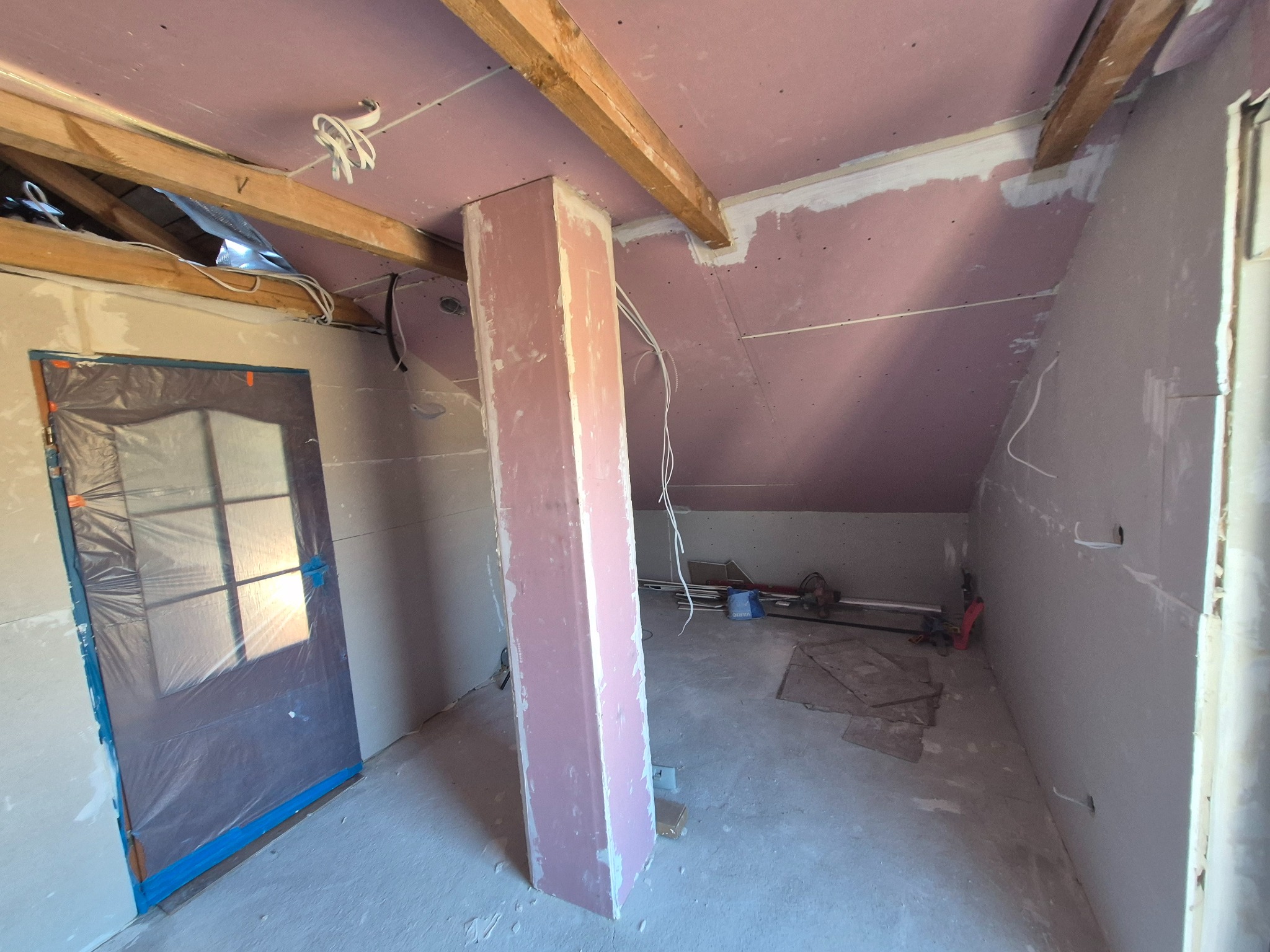 Poddasze w trakcie wykańczania: różowe płyty gipsowe na skosach, drewniane belki stropowe, zabezpieczone drzwi folią, pionowy słup obłożony płytami, widoczne przewody elektryczne.