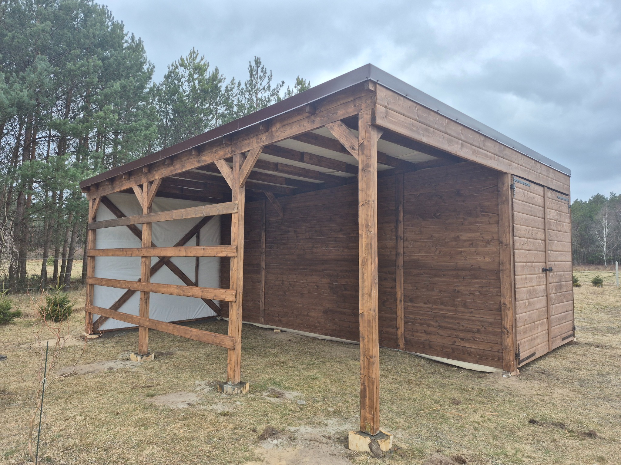 Drewniana wiata z brązowym dachem na zewnątrz, z widoczną konstrukcją i białym materiałem w tle. Widoczne drzewa i trawa.