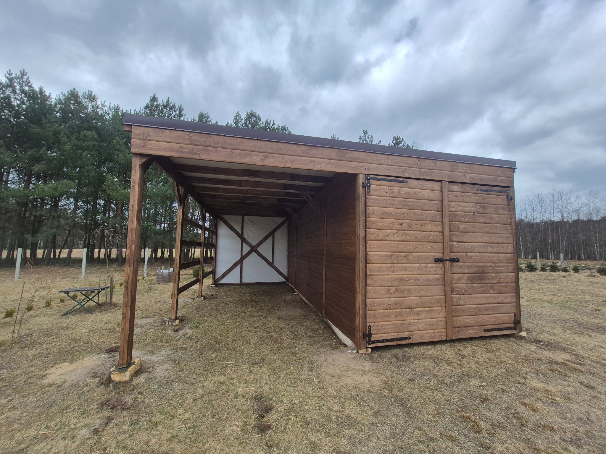 Drewniana wiata z zamykaną częścią, wykonana z desek w odcieniach brązu, na tle drzew i pochmurnego nieba. Widoczna konstrukcja wsporcza i zadaszenie.