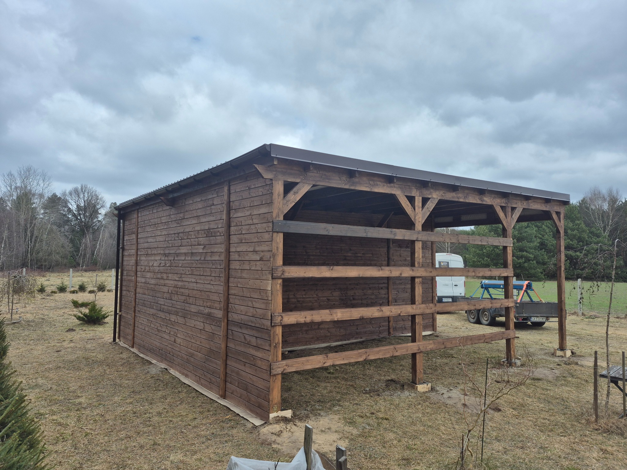 Drewniana konstrukcja zadaszona brązowym blachodachówką, częściowo zabudowana deskami, na trawiastym terenie. W tle drzewa i pochmurne niebo.
