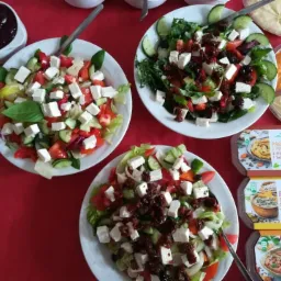 Trzy talerze sałatek z serem feta, pomidorami, ogórkami i różnymi dodatkami na czerwonym obrusie, obok dipy i dżem w białych naczyniach, oraz pudełka z pastami.