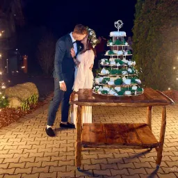 Pocałunek pary młodej przy tortowej piramidzie na rustykalnym stole, otoczonej fontannami iskier na tle nocnego ogrodu. Dekoracje weselne.