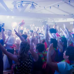 Rozbawiony tłum gości weselnych z podniesionymi rękami, w tle kamerzysta rejestrujący wydarzenie, dekoracje świetlne i kolorowe rekwizyty.