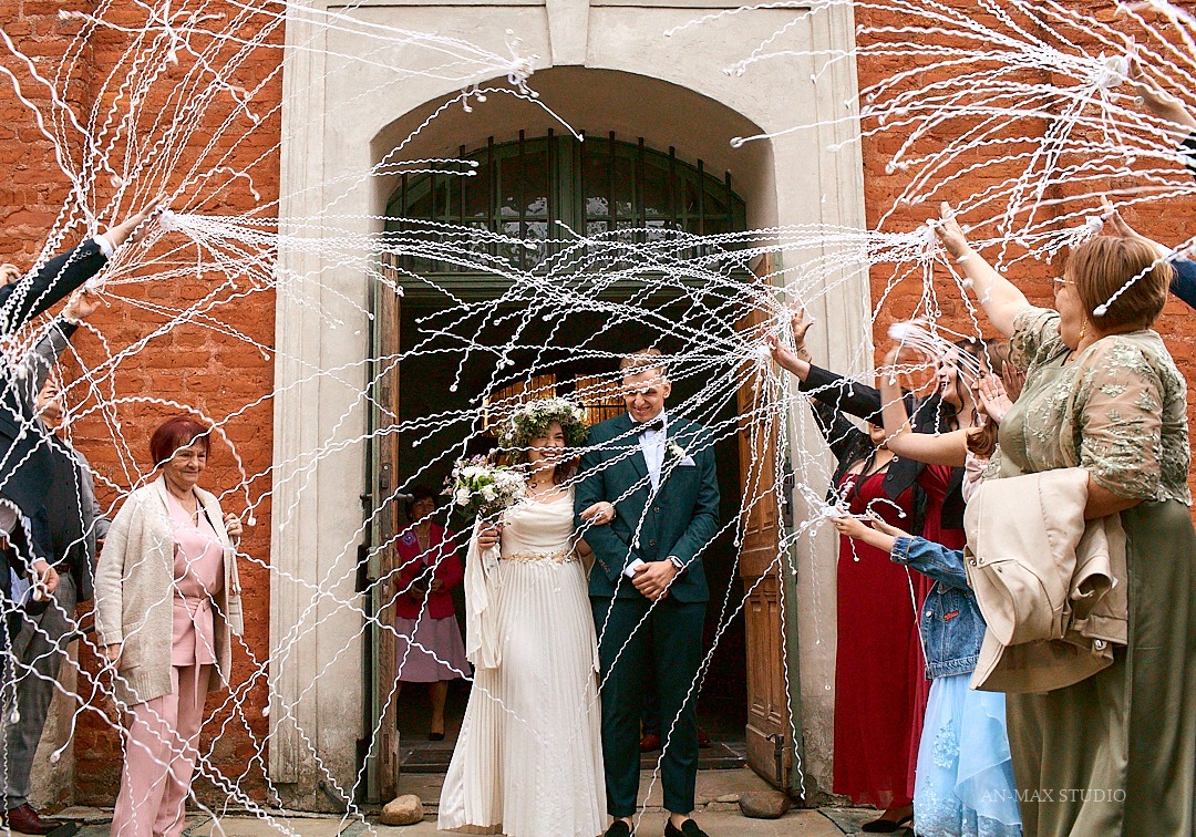 Młoda para wychodzi z kościoła, obsypywana serpentynami przez gości. Panna młoda w wianku i sukni, pan młody w zielonym garniturze. Czerwona cegła kościoła w tle.