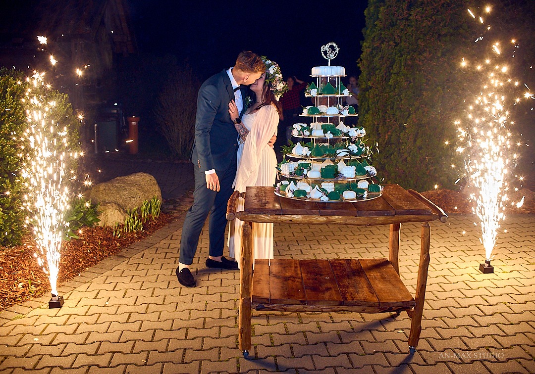 Pocałunek pary młodej przy tortowej piramidzie na rustykalnym stole, otoczonej fontannami iskier na tle nocnego ogrodu. Dekoracje weselne.