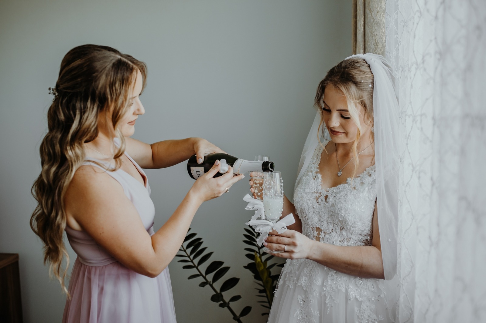 Panna młoda w koronkowej sukni z welonem, podczas przygotowań, nalewa szampana do kieliszka. Obok stoi druhna w jasnej sukience. W tle roślina doniczkowa i okno.