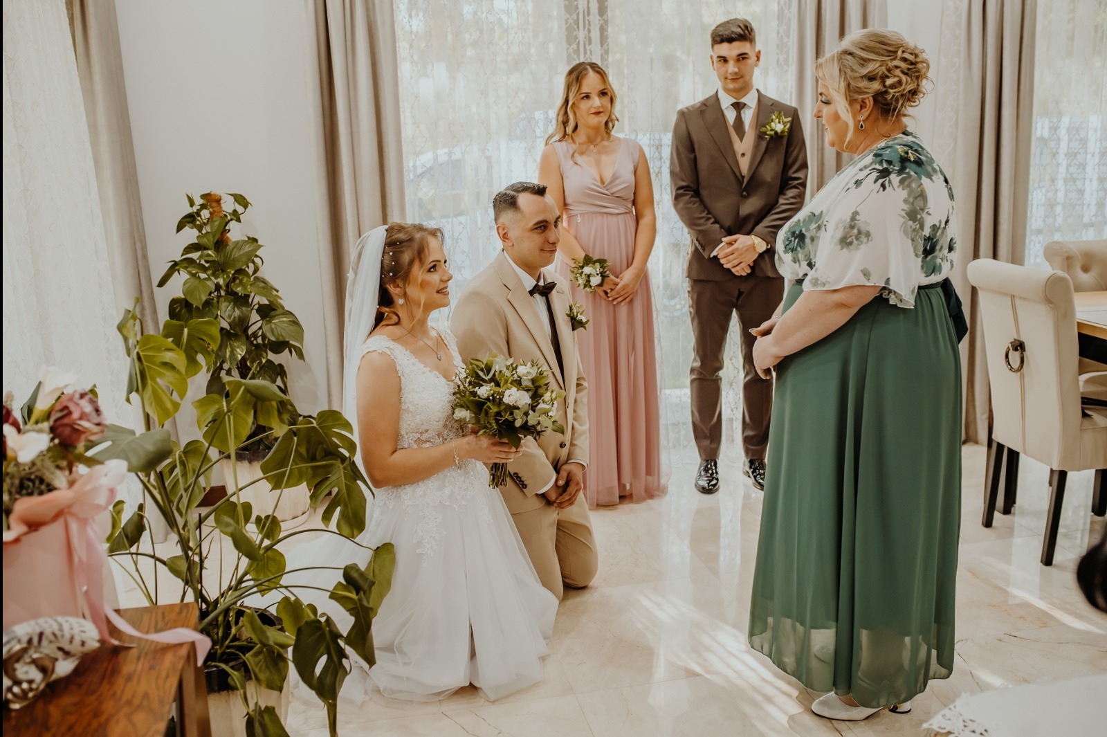 Uroczysta chwila: Pan młody klęczy przed panną młodą z bukietem, w tle świadkowie i kobieta w zielonej sukni, jasne wnętrze z roślinami. Romantyczna atmosfera.