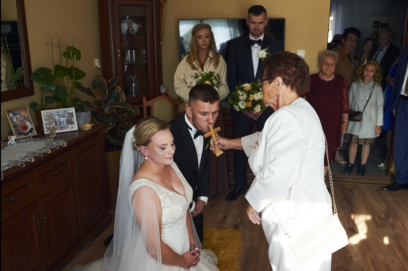 Błogosławieństwo krzyżem podczas ceremonii ślubnej w domu, z parą młodą klęczącą na dywanie i gośćmi w tle. Ujęcie z bliska, ciepłe oświetlenie.