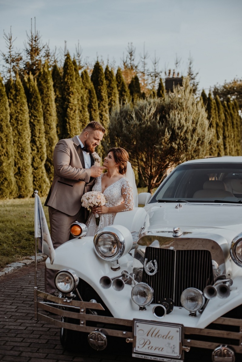 Uśmiechnięta para młoda w eleganckich strojach pozuje przy białym, luksusowym samochodzie z tabliczką 'Młoda Para' na tle zielonych drzew.