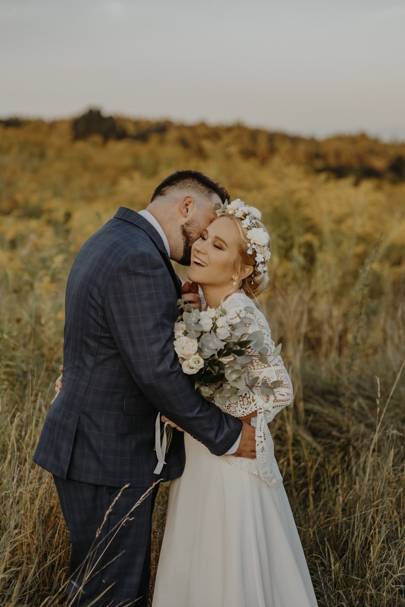 Uśmiechnięta panna młoda w wianku i koronkowej sukni, obejmowana przez pana młodego w kraciastym garniturze na tle łąki w ciepłym świetle.