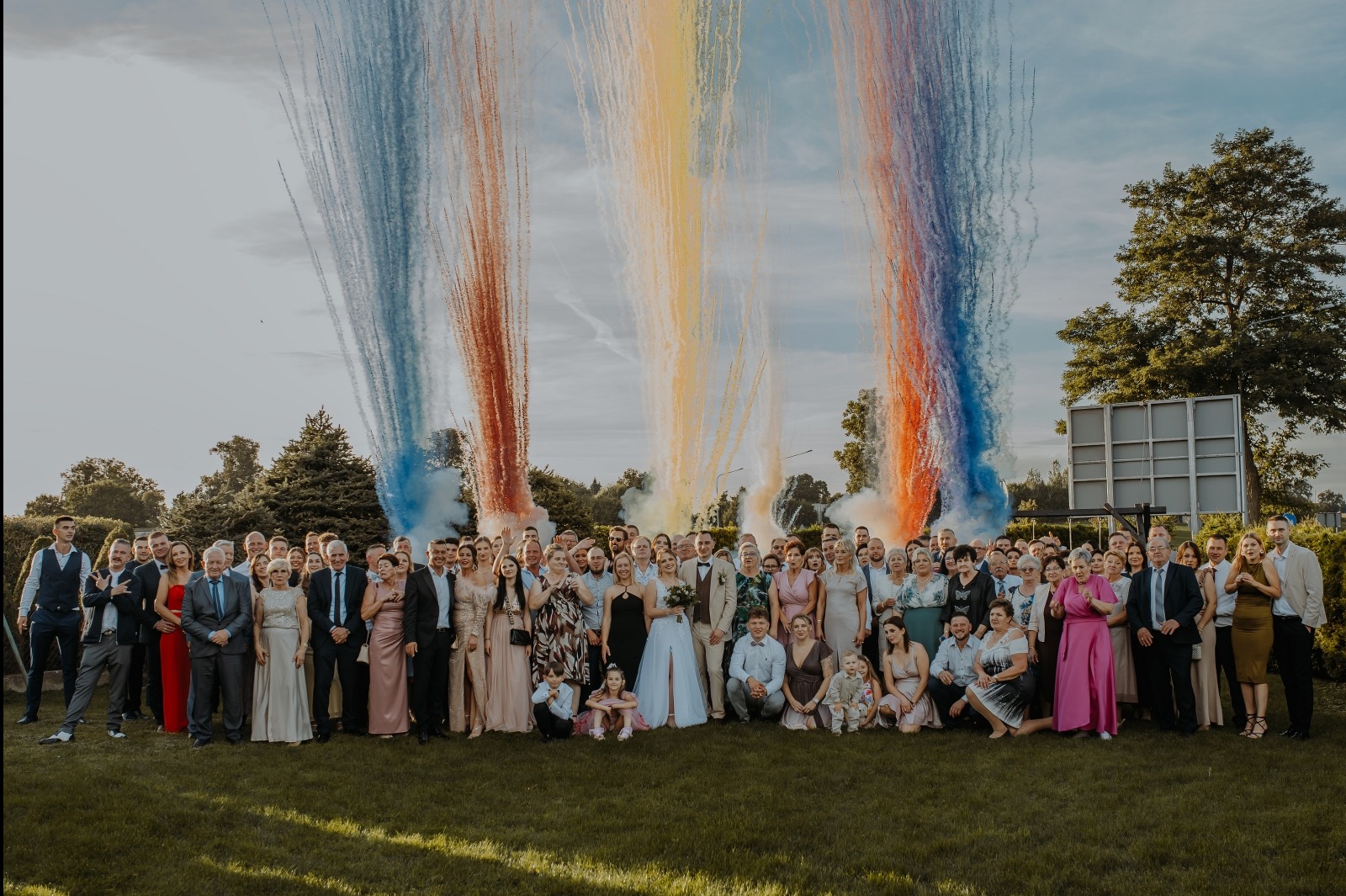 Grupowe zdjęcie ślubne z gośćmi na tle kolorowych fontann pirotechnicznych, wykonane z niskiego kąta, ukazujące radość i świętowanie.