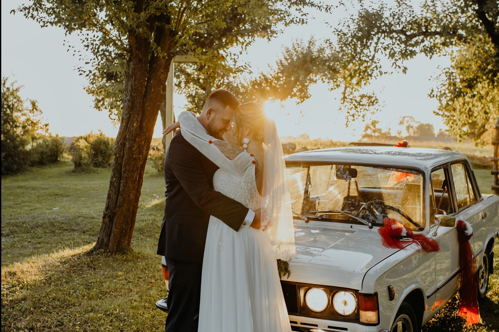 Para młoda w objęciach na tle białego, klasycznego samochodu udekorowanego czerwonymi wstążkami, w słoneczny dzień. Romantyczna sesja ślubna w plenerze.