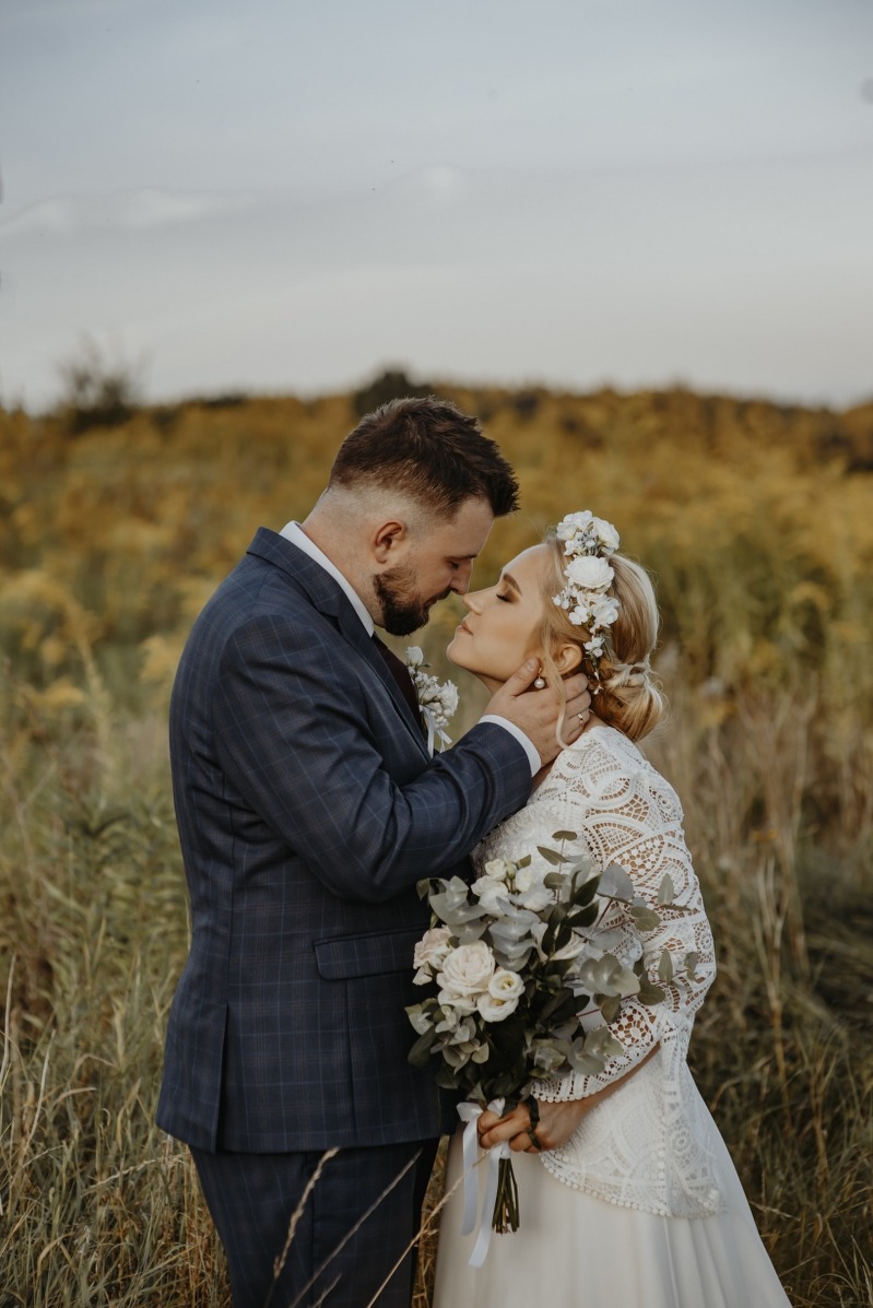 Para młoda w plenerze: panna młoda w koronkowej sukni i wianku, pan młody w garniturze w kratę, delikatny bukiet, subtelne oświetlenie, naturalne tło łąki.