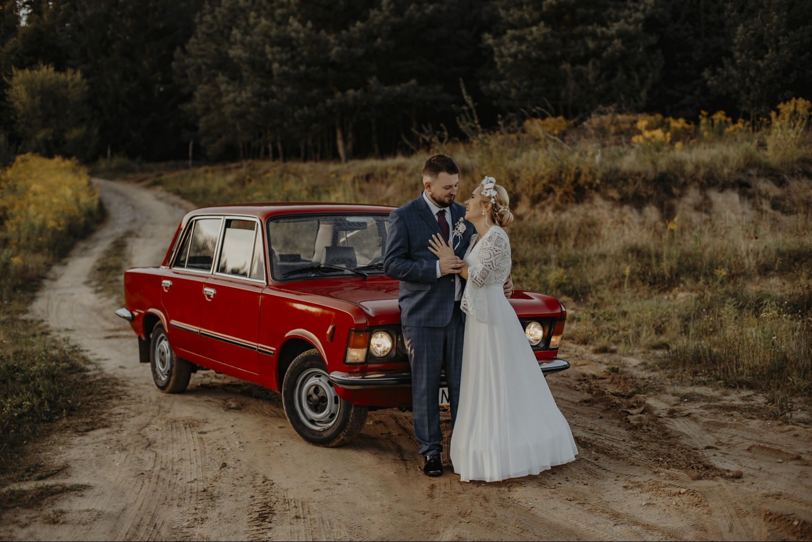 Para młoda w eleganckich strojach pozuje przy czerwonym Fiacie 125p na polnej drodze, w tle las. Romantyczna sesja ślubna w stylu vintage.