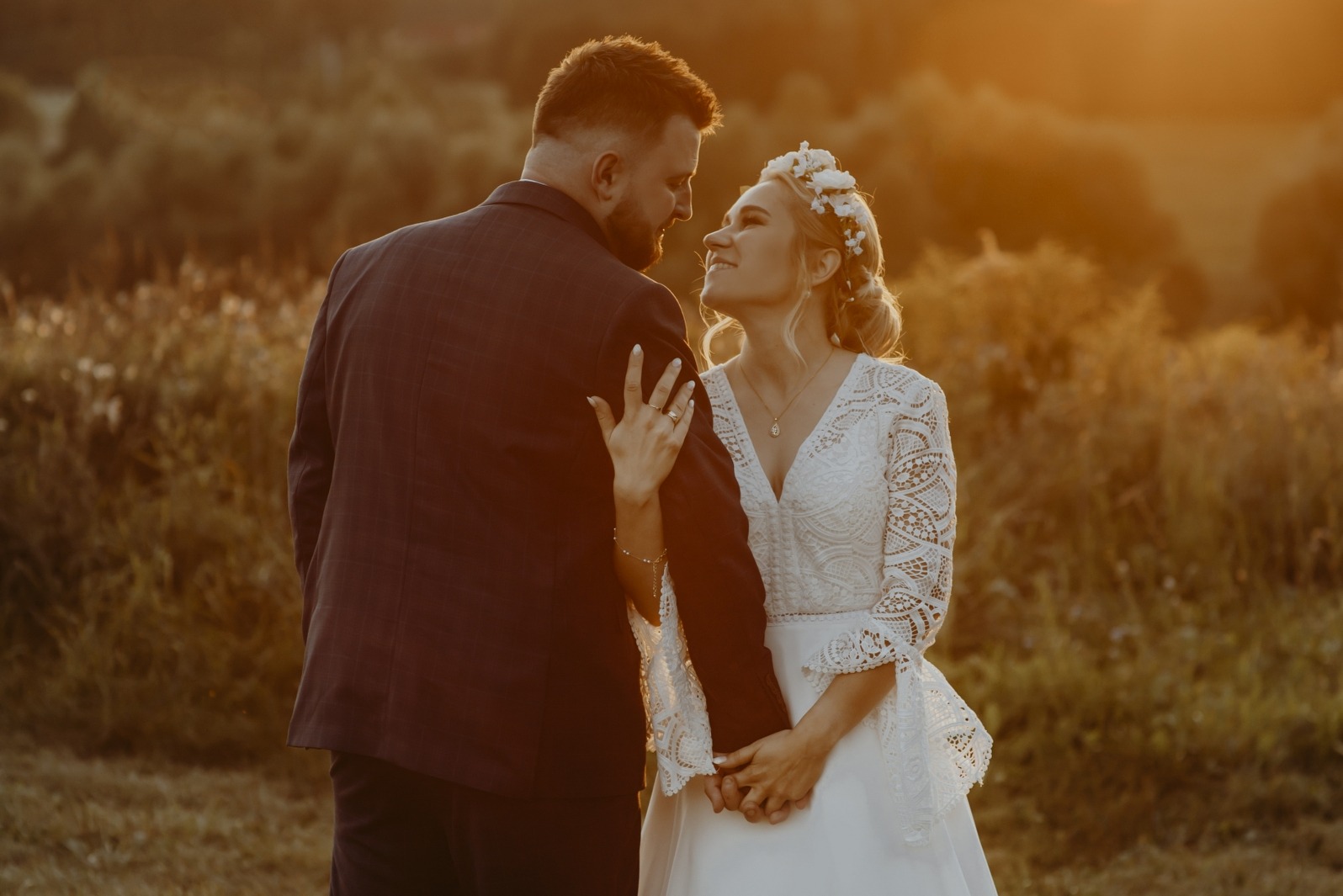 Para młoda w plenerze o zachodzie słońca. Panna młoda w koronkowej sukni i wianku spogląda na pana młodego w bordowym garniturze. Romantyczna atmosfera.