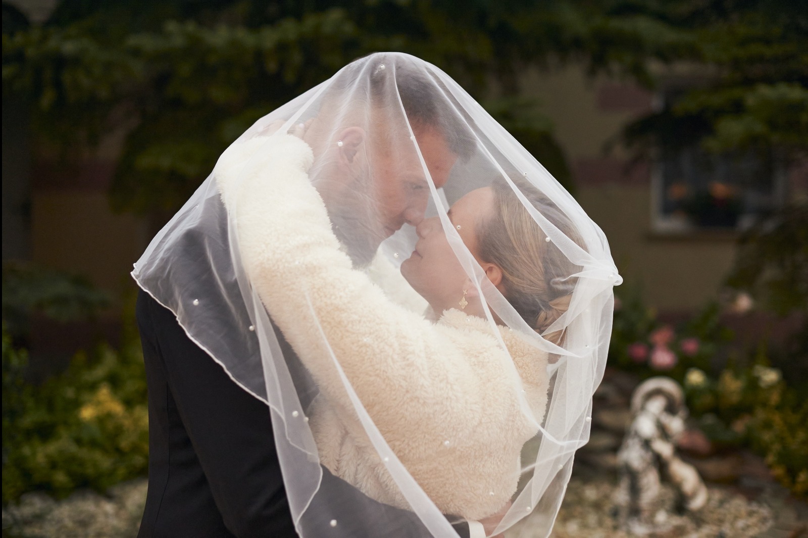 Para młoda w objęciach pod delikatnym, perłowym welonem na tle zieleni. Romantyczny moment uchwycony z bliska, z subtelnym oświetleniem.