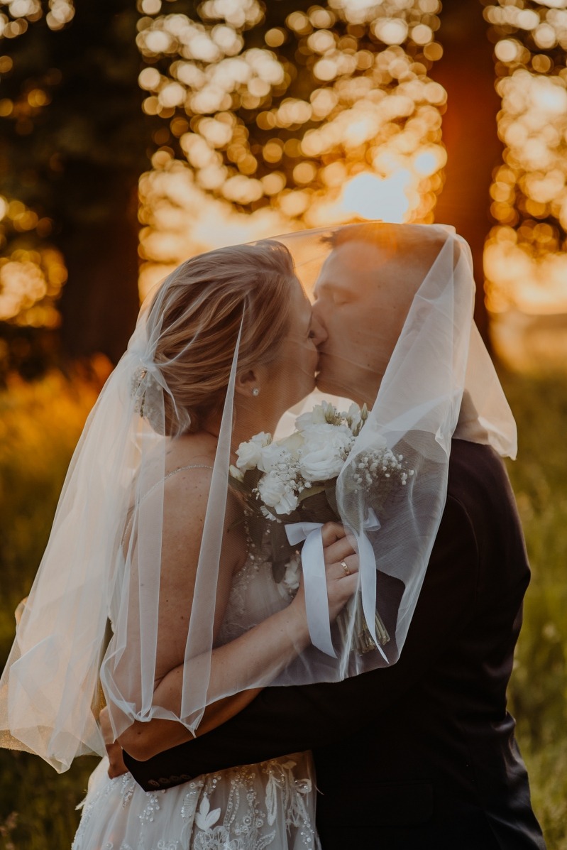 Pocałunek młodej pary pod welonem na tle zachodzącego słońca, bukiet białych kwiatów w dłoniach panny młodej, pierścionek na palcu. Romantyczna fotografia ślubna w plenerze.