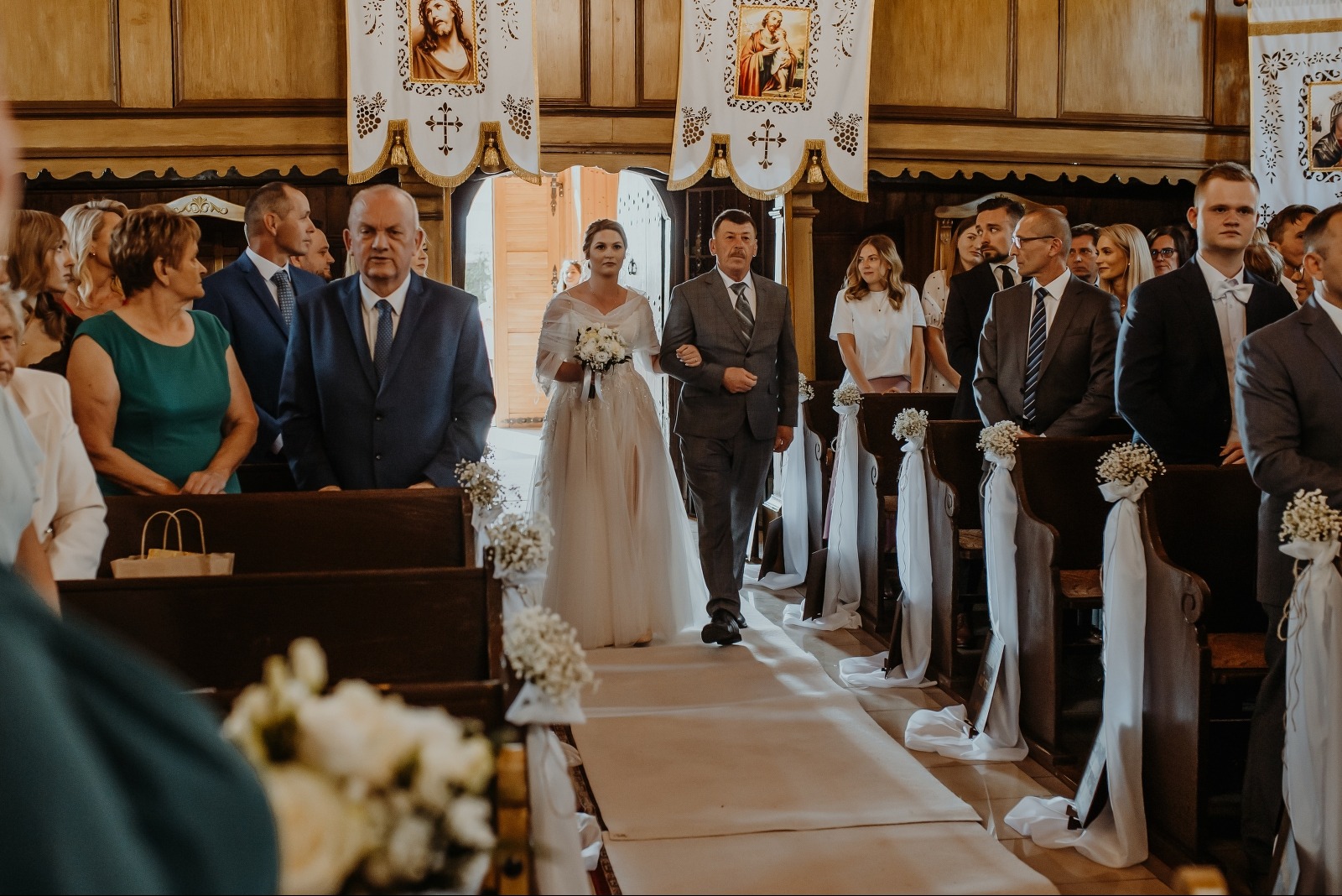 Panna młoda w sukni ecru prowadzona do ołtarza przez ojca w szarym garniturze, w kościele z dekoracjami kwiatowymi i białymi wstążkami na ławkach.