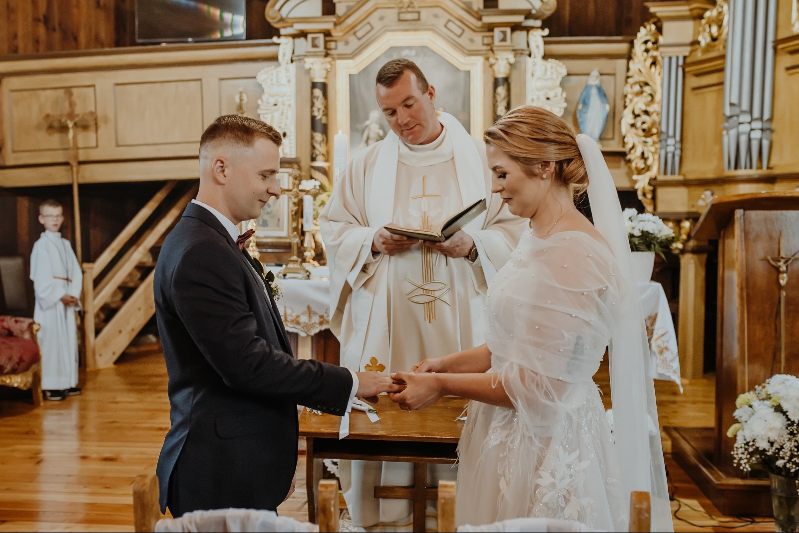Wymiana obrączek podczas ceremonii ślubnej w kościele w Olsztynie. Para młoda, ksiądz i ministrant na tle drewnianego ołtarza. Ujęcie z bliska, ciepłe oświetlenie.