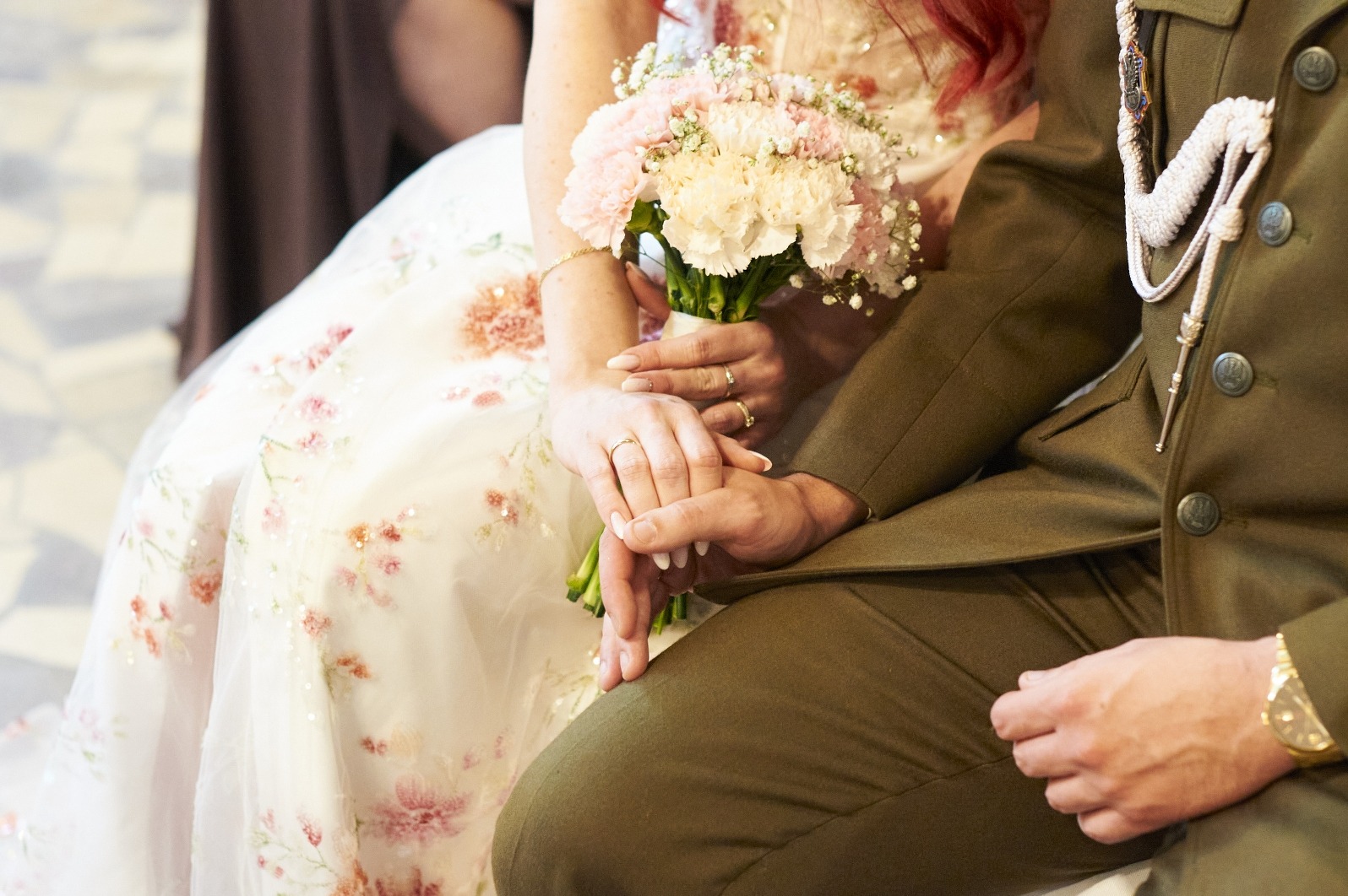 Dłonie pary młodej z obrączkami, panna młoda trzyma bukiet, pan młody w mundurze. Ujęcie z bliska, skupione na dłoniach i detalach stroju.