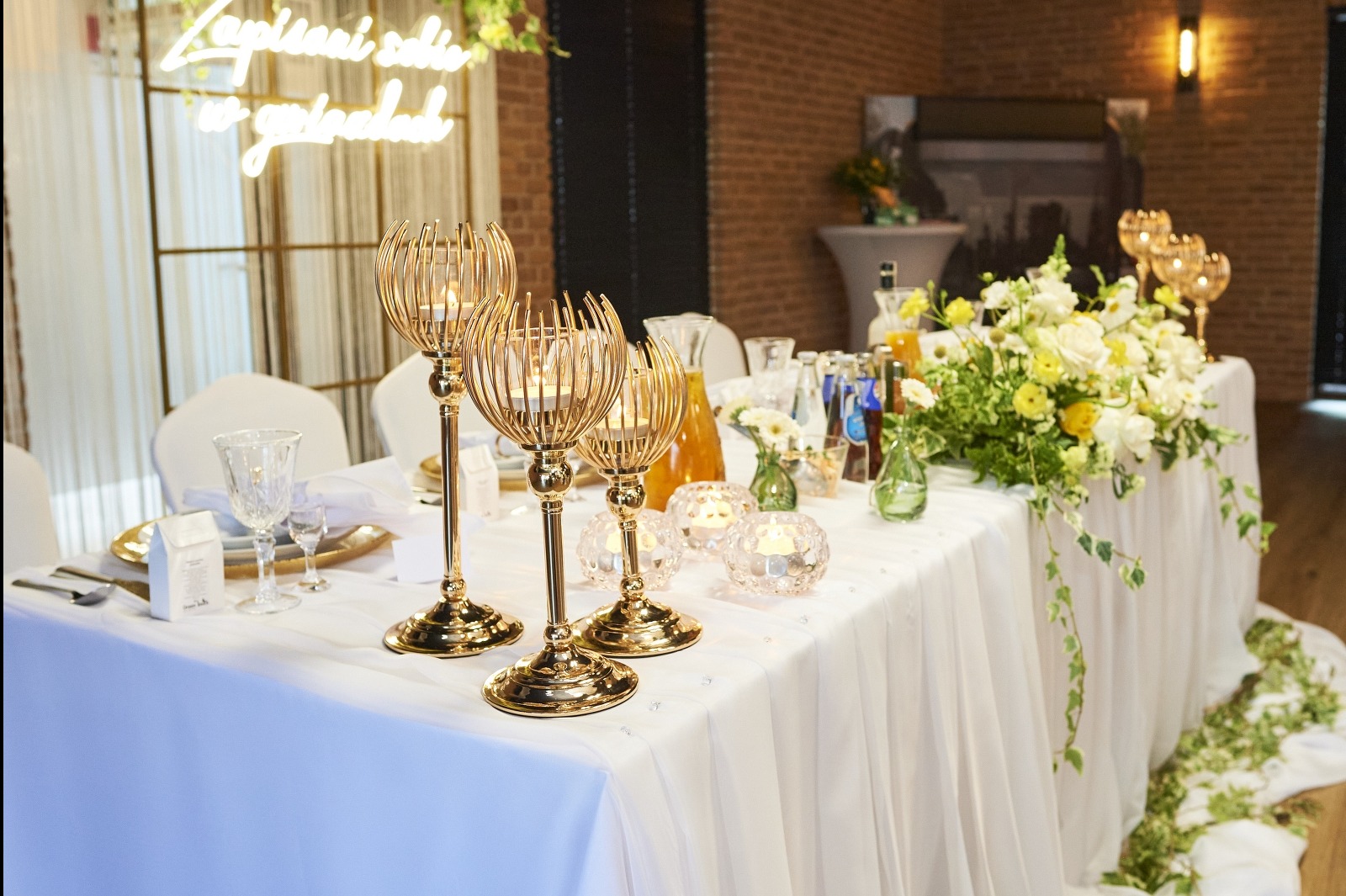 Uroczysty stół weselny z białym obrusem, złotymi świecznikami, kwiatami i napojami. W tle neonowy napis i ceglana ściana.