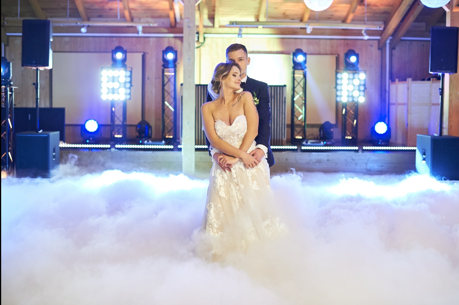 Para młoda w objęciach na parkiecie w chmurach, oświetlonym niebieskimi światłami. Panna młoda w koronkowej sukni, pan młody w granatowym garniturze. Romantyczna atmosfera.