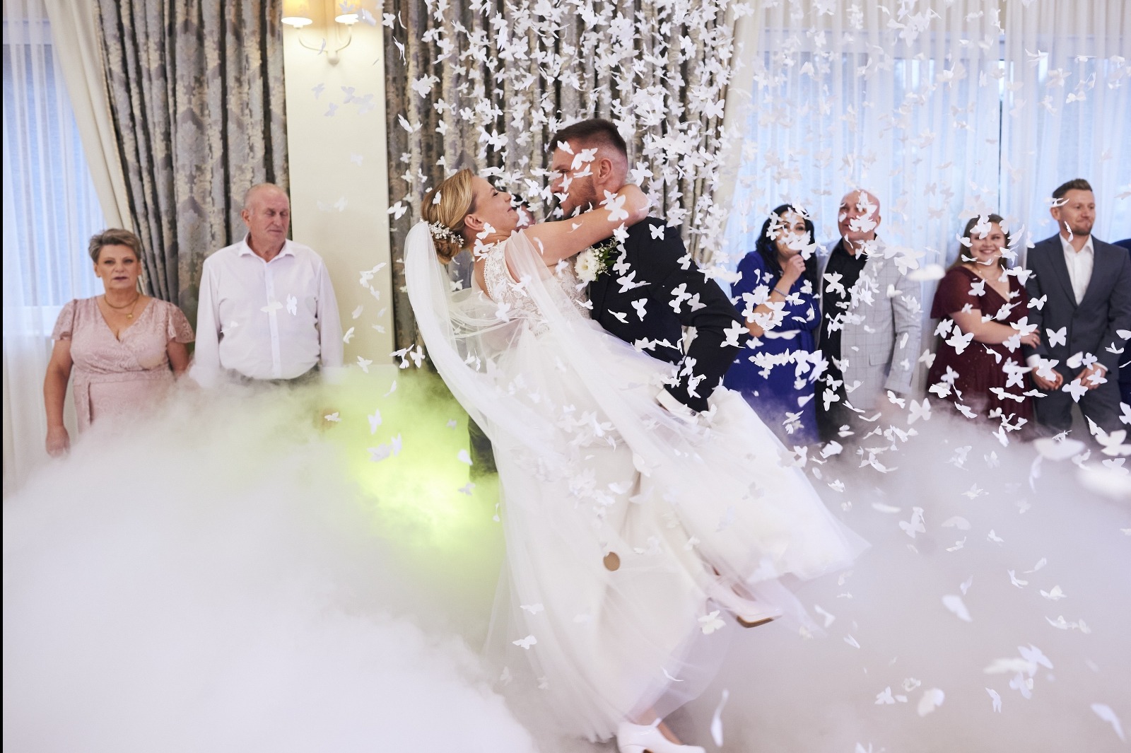 Uśmiechnięta para młoda w tańcu, unoszona przez pana młodego, otoczona białymi konfetti i dymem na tle gości weselnych i dekoracji sali.