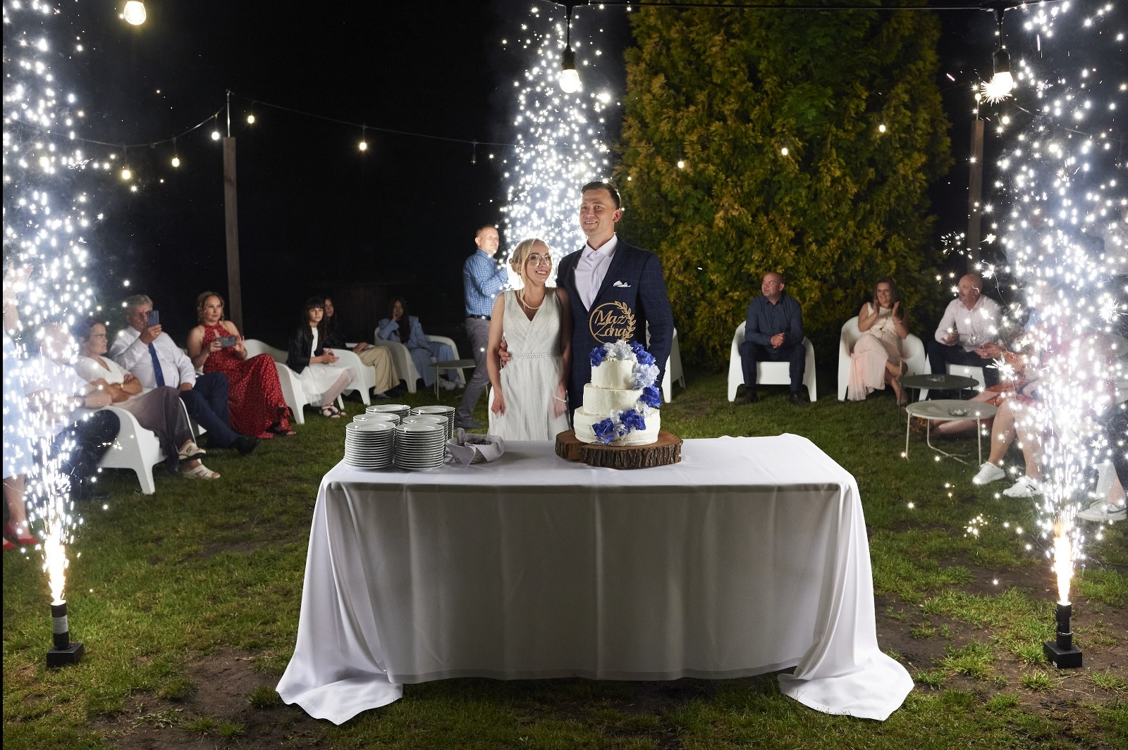 Uśmiechnięta para młoda stoi za stołem z tortem weselnym, otoczona gośćmi i fontannami iskier na tle nocnego ogrodu z girlandami świateł.
