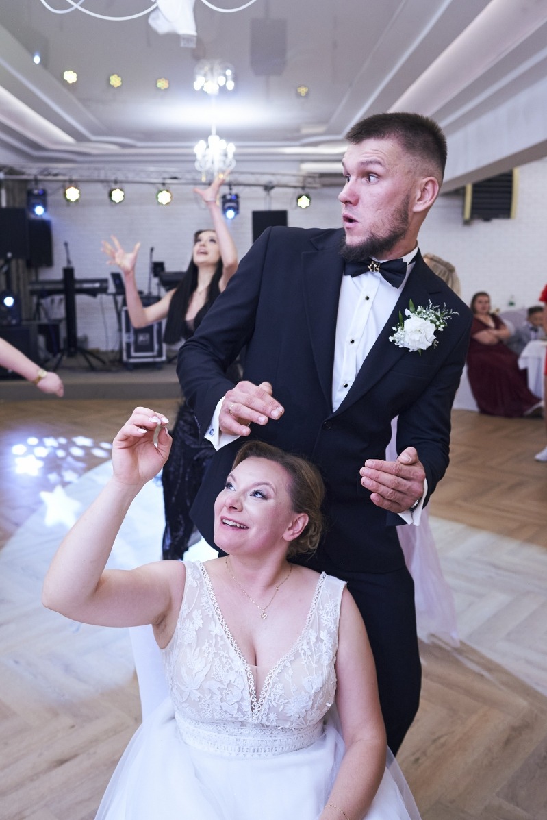 Uśmiechnięta panna młoda łapie welon rzucony przez druhnę, obok stoi zaskoczony pan młody w smokingu. W tle sala balowa z oświetleniem.