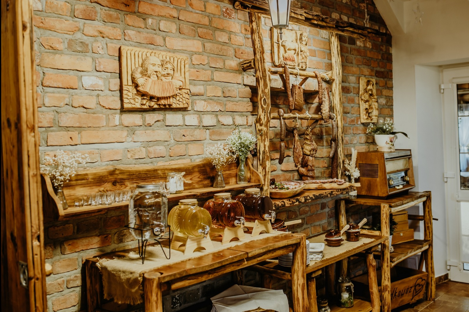 Rustykalny stół z napojami w szklanych słojach i wędlinami na drewnianej drabinie, ceglana ściana z rzeźbami, dekoracje kwiatowe, radio w stylu vintage.