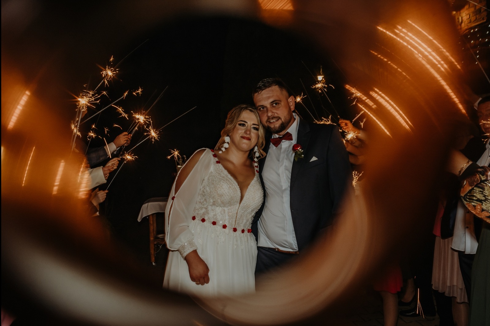 Uśmiechnięta para młoda w otoczeniu gości z zimnymi ogniami, uchwycona przez unikalny, rozmyty kadr, tworzący efektowne obramowanie.