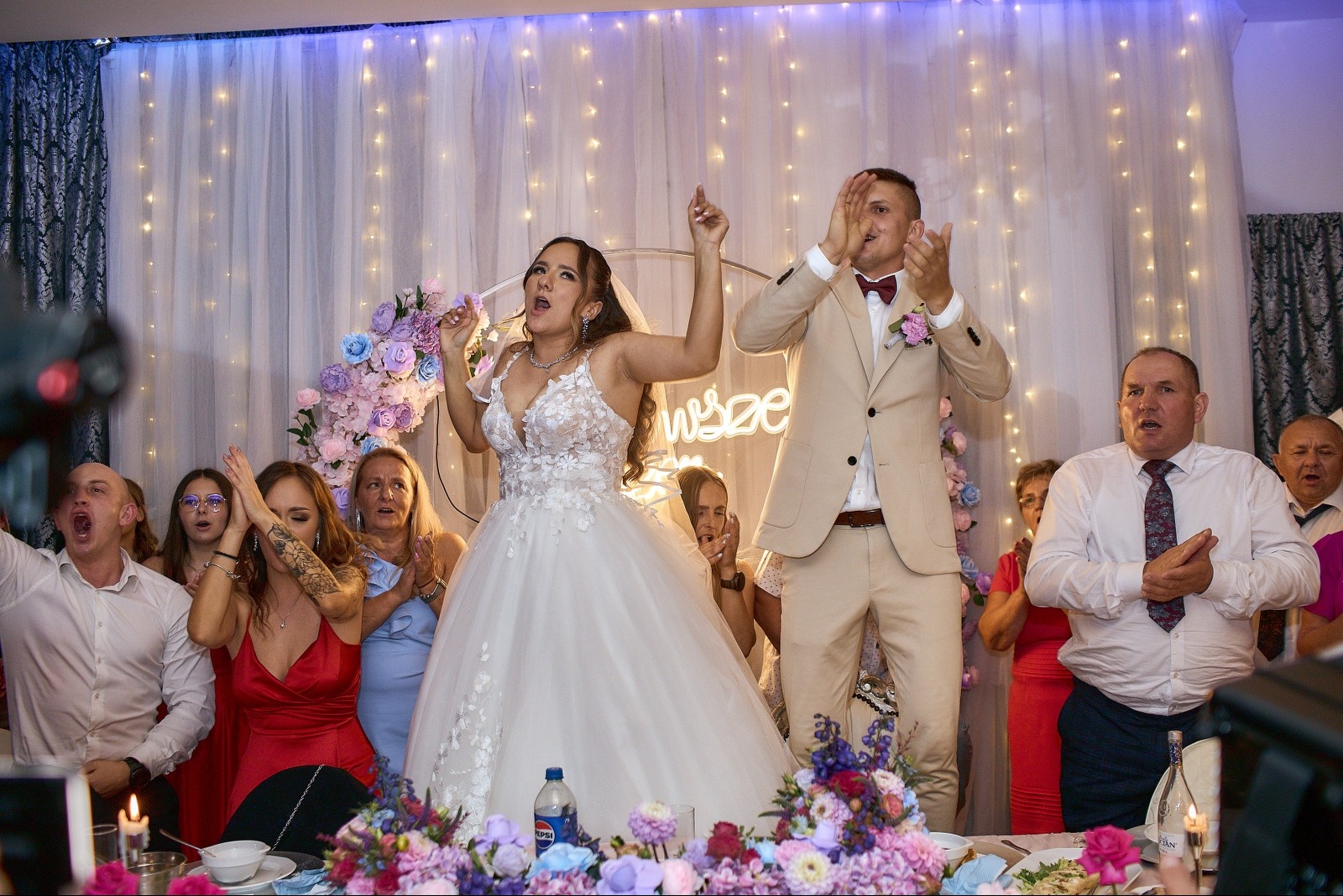 Radosna para młoda w tańcu podczas wesela, otoczona gośćmi i dekoracjami kwiatowymi. W tle kurtyna ze światełek i neonowy napis.