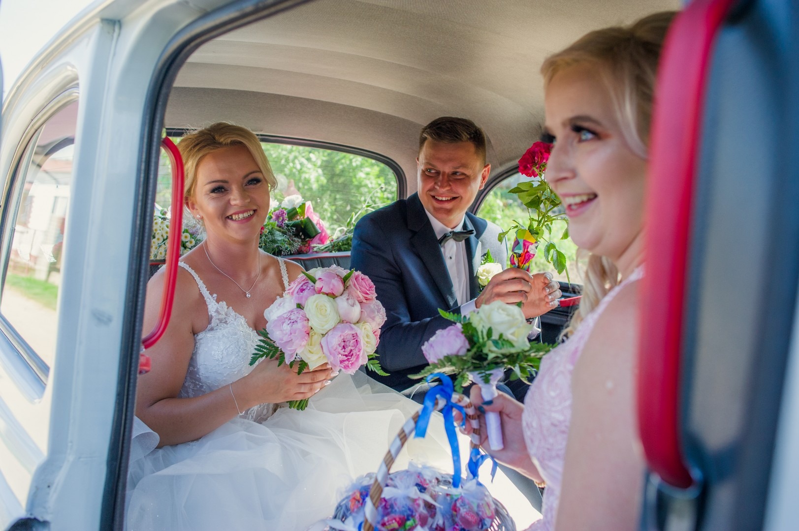 Panna młoda w białej sukni z bukietem różowych i białych piwonii, uśmiechnięty pan młody w ciemnym garniturze i muszce w tle, druhna z koszykiem kwiatów i różyczką w dłoni w samochodzie w drodze...