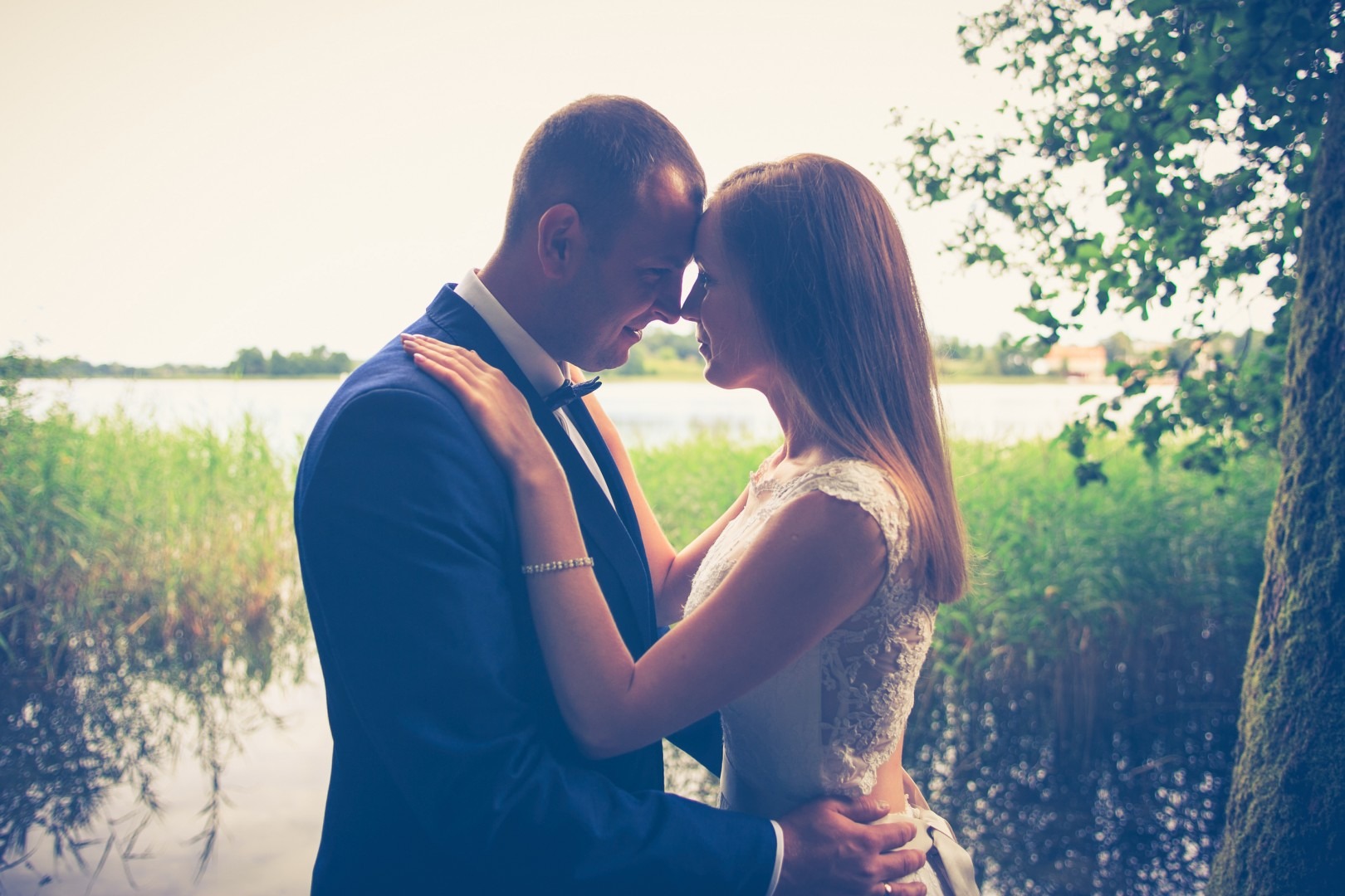 Para młoda, on w granatowym garniturze z muszką, ona w koronkowej sukni, stykają się czołami na tle jeziora i trzcin.