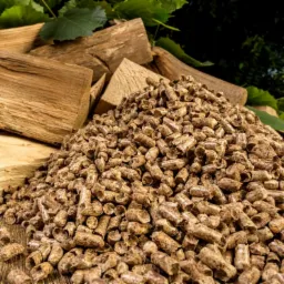 Góra jasnobrązowego pelletu drzewnego na drewnianym blacie, z kawałkami drewna w tle i zielonymi liśćmi w górnej części kadru.