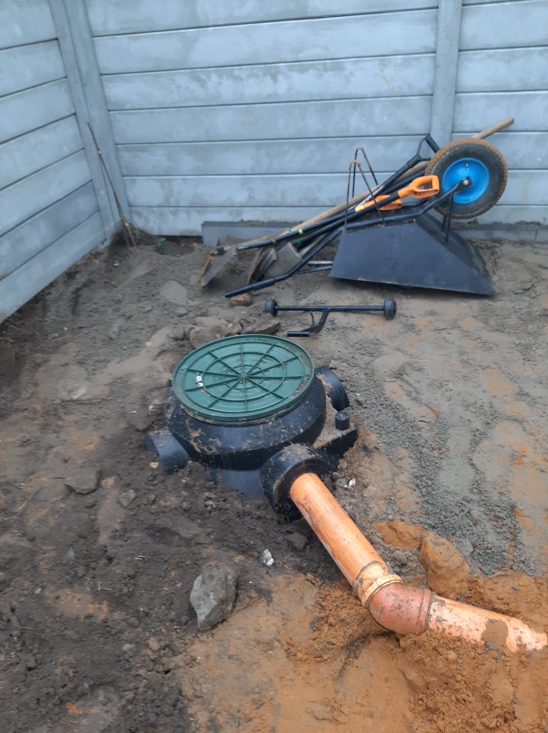 Montaż studzienki kanalizacyjnej z rurą odpływową w wykopie, widoczne narzędzia i taczka w tle, szara betonowa ściana w tle.