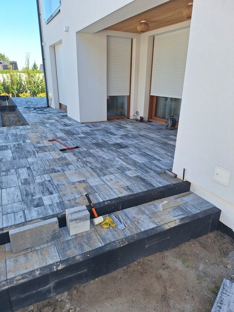 Układanie kostki brukowej przed nowoczesnym domem z białymi ścianami i drewnianymi elementami, widoczne narzędzia pracy, etap wykańczania schodów z kostki.