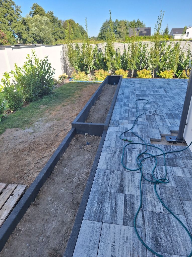 Fragment tarasu z szarej kostki, widoczny wąż ogrodowy i przygotowane miejsce na rabatę kwiatową z obrzeżami, w tle biały mur i zieleń.