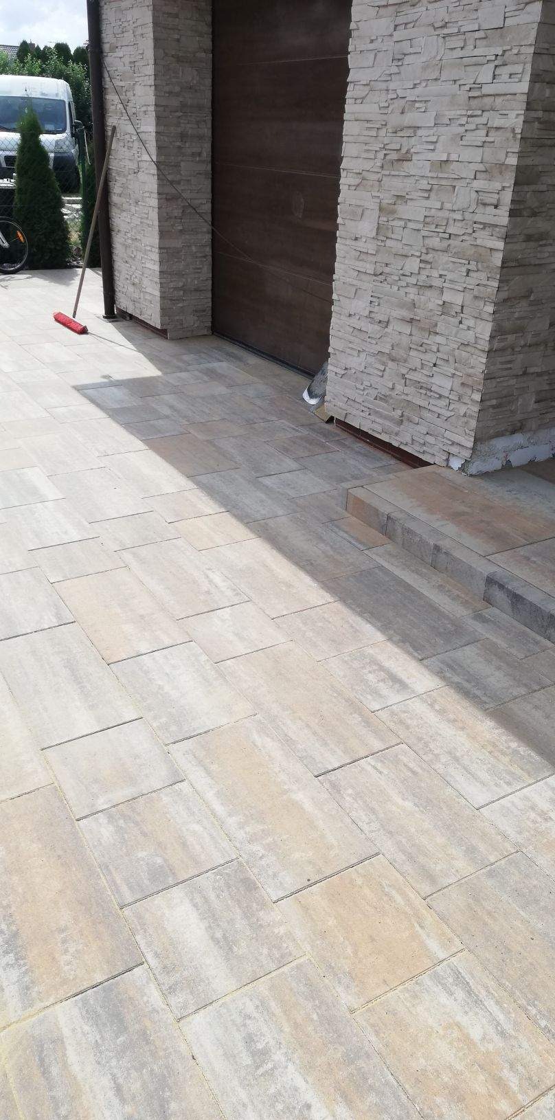 Ułożona beżowo-szara kostka brukowa przed garażem z kamienną elewacją, widoczna czerwona zmiotka oparta o ścianę.