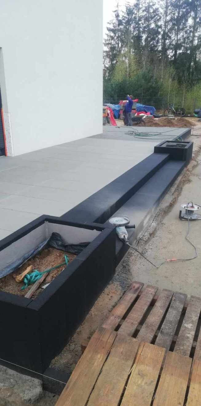 Betonowe donice z czarnym wykończeniem, częściowo wypełnione ziemią, z widocznymi narzędziami budowlanymi i paletą w tle, na tle świeżo ułożonej betonowej kostki i białego budynku.