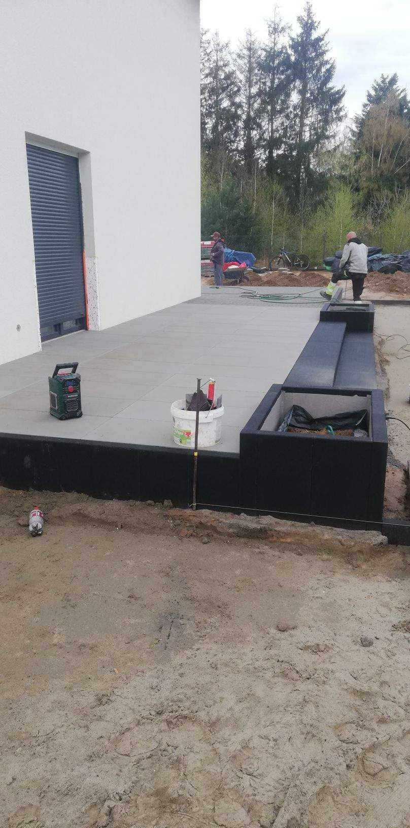 Układanie szarej kostki brukowej na tarasie przy domu, widoczne narzędzia i pracownicy podczas montażu obrzeży rabat